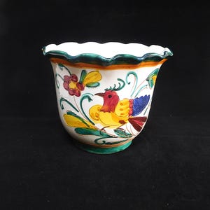 May include: A white ceramic planter with a scalloped rim, featuring a hand-painted design of a colorful bird and floral elements. The bird has red, yellow, and blue feathers. The planter has a green base and rim, and is suitable for indoor or outdoor use.
