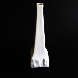 May include: A white ceramic vase with a textured base. The vase is tall and slender, with a square opening at the top.