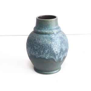 Puede incluir: Un jarrón de cerámica azul verdoso y azul con un cuerpo redondeado y un cuello estrecho. El jarrón tiene un aspecto texturizado y moteado con un borde oscuro. La base es un tono más oscuro de azul verdoso. Una pieza decorativa para el hogar.