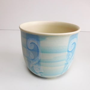 Puede incluir: Un cuenco de cerámica con interior crema claro y exterior azul claro. El exterior presenta rayas horizontales y un diseño decorativo en espiral en un tono más oscuro de azul. El cuenco tiene forma redondeada y un acabado liso.