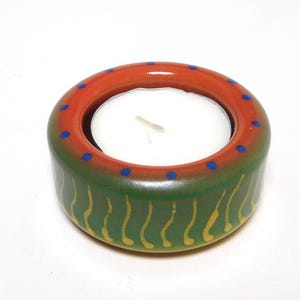 May include: A ceramic tealight candle holder. The holder is round with a red rim dotted with blue circles. The base is green with yellow curved lines. A white candle sits inside the holder.