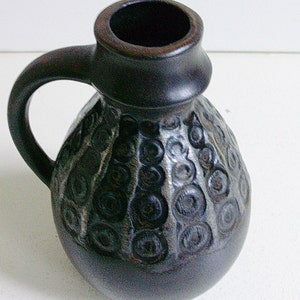 May include: A dark brown ceramic pitcher with a textured surface and a circular pattern. The pitcher has a handle and a wide opening.