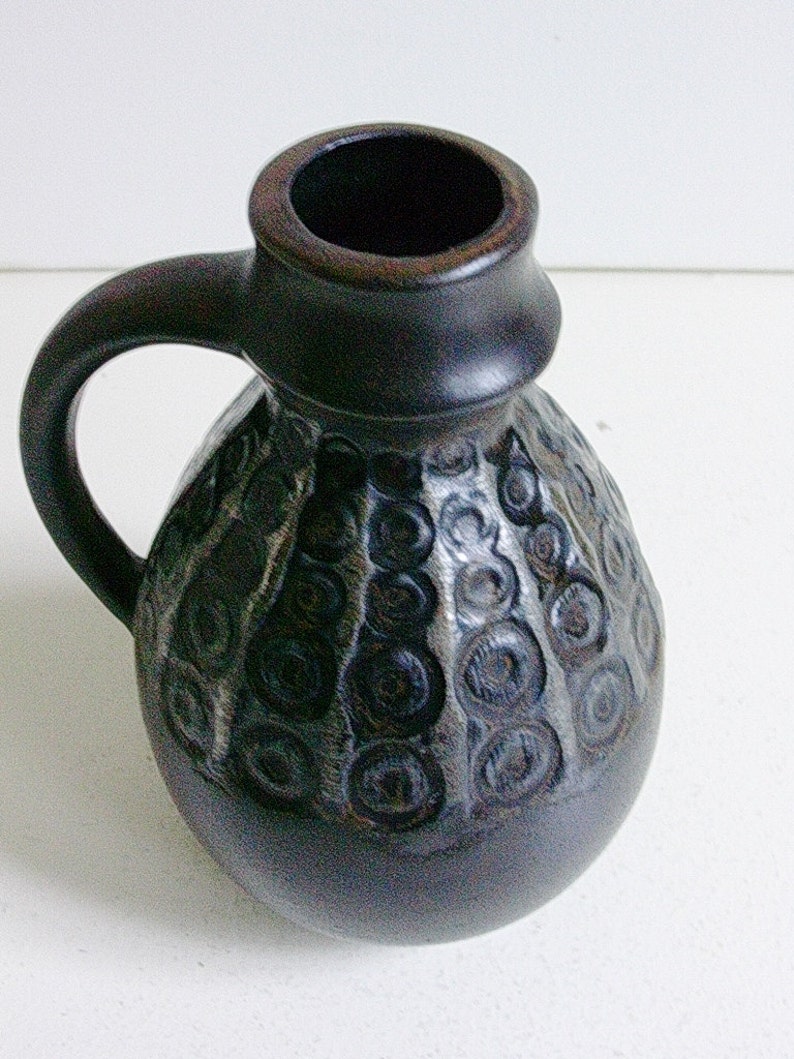May include: A dark brown ceramic pitcher with a textured surface and a circular pattern. The pitcher has a handle and a wide opening.