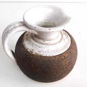 Può includere: Brocca in ceramica fatta a mano con base marrone rotonda e strutturata e parte superiore bianca svasata. Il manico e il bordo della brocca sono bianchi. La brocca è un pezzo unico di ceramica.