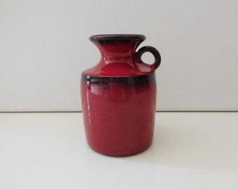 May include: A small red ceramic vase with a black rim and a handle. The vase has a rounded body and a narrow neck.