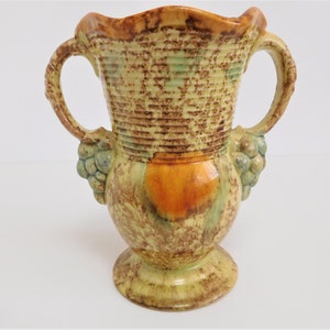 以下が含まれることがあります： 2つの取っ手がついたビンテージの陶器の花瓶。茶色、緑、黄色のまだら模様の釉薬が施されています。取っ手はブドウの房で飾られています。