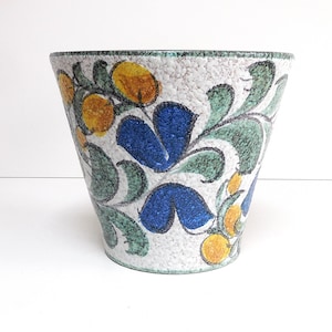 Peut inclure: Un pot de fleurs en céramique conique avec une base blanche texturée ornée d'un motif botanique peint à la main. Le design présente des fruits circulaires jaune vif, des pétales de fleurs bleu profond et des vignes vertes feuillues.