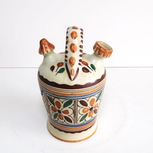 Może przedstawiać: Ceramiczny dzbanek na wodę w kremowym kolorze, z uchwytem i dwoma wylewkami. Dzbanek ma brązowe, pomarańczowe, niebieskie i zielone wzory kwiatowe i geometryczne. Uchwyt ma serię pomarańczowych kropek.