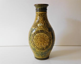 May include: A ceramic vase with a glossy mustard yellow and dark brown glaze. The body features a raised relief pattern of a central mandala surrounded by swirling motifs. A small white sticker with the text JAV is visible on the upper neck.