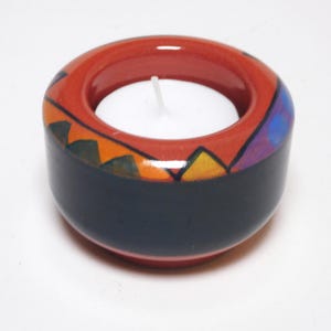May include: A ceramic candle holder with a red rim and a dark blue base. The rim is decorated with colorful geometric patterns in orange, green, and purple. A white tea light candle sits inside the holder. The candle holder is a decorative item.