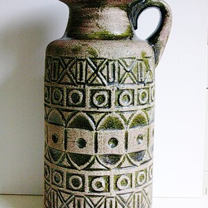May include: A tall, green and brown ceramic vase with a geometric pattern. The vase has a wide mouth and a handle.