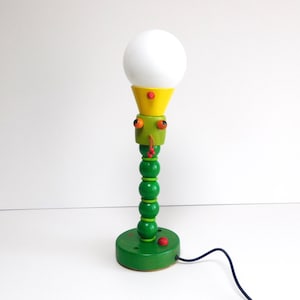 Può includere: Una lampada da tavolo stravagante, fatta a mano, con una lampadina bianca a forma di globo. La lampada presenta una base e uno stelo in legno verde, una testa gialla e verde e dettagli rossi. È collegata a un cavo di alimentazione nero. La lampada è alta circa 30 cm.