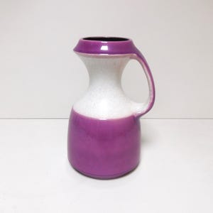 May include: A ceramic pitcher with a handle, featuring a two-tone design. The base and rim are a vibrant purple, while the body is white with a speckled texture. The interior is black.