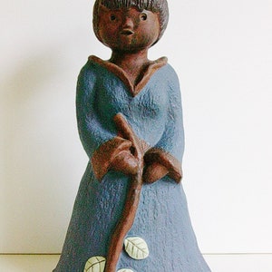 May include: A ceramic figurine of a person wearing a blue robe and holding a stick. The figurine has brown hair and a brown face. The robe has white leaf accents.