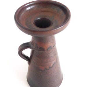Può includere: Un vaso in ceramica marrone con una parte superiore ampia e poco profonda e un manico. Il vaso ha un corpo conico e un interno marrone scuro. L'esterno ha un aspetto strutturato e terroso, con variazioni di colore. Il vaso è probabilmente utilizzato per scopi decorativi.