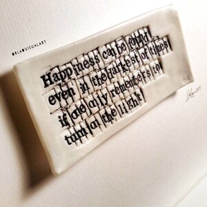 May include: A rectangular, off-white ceramic plaque with the quote "Happiness can be found even in the darkest of times if one only remembers to turn on the light" in black, typewriter-style font. The plaque is slightly raised and has a textured surface.