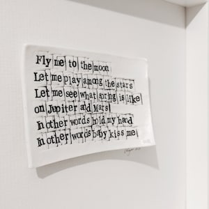 May include: A white ceramic tile with black text that reads "Fly me to the moon Let me play among the stars Let me see what spring is like on Jupiter and Mars In other words hold my hand In other words baby kiss me". The text is in a mosaic style, with each word made up of individual tiles.