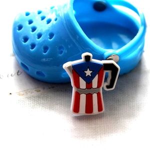 May include: A blue clog shoe with a coffee pot charm. The charm is a miniature replica of a coffee pot, featuring the colors of the Puerto Rican flag: red, white, and blue. The charm has a black handle.