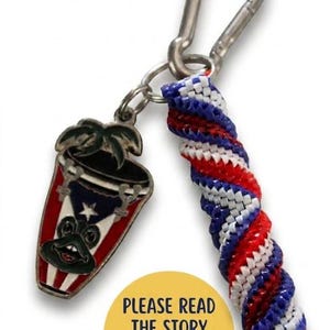 May include: A keychain featuring a silver carabiner, a metal charm shaped like a conga drum with a frog face and a Puerto Rican flag design, and a twisted cord in red, white, and blue. A yellow circular sticker reads PLEASE READ THE STORY BEHIND THIS KEYCHAIN.