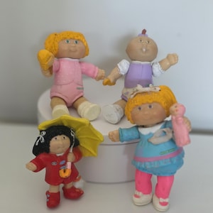 May include: Four small plastic dolls with round faces and yarn-like hair. One wears a pink outfit, one a purple and white outfit, one a red coat with a yellow umbrella, and one a blue dress with pink pants, holding a tiny pink doll figure.