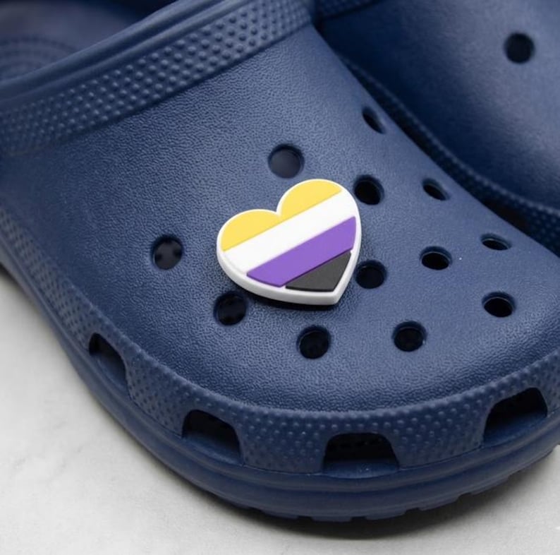 A heart-shaped soft PVC shoe charm featuring the non-binary pride flag with horizontal stripes of yellow, white, purple, and black, resting on a grey texture background.