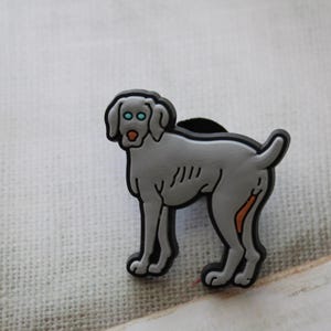 May include: A gray Weimaraner dog charm with blue eyes and an orange nose. The charm is made of rubber and has a black base.