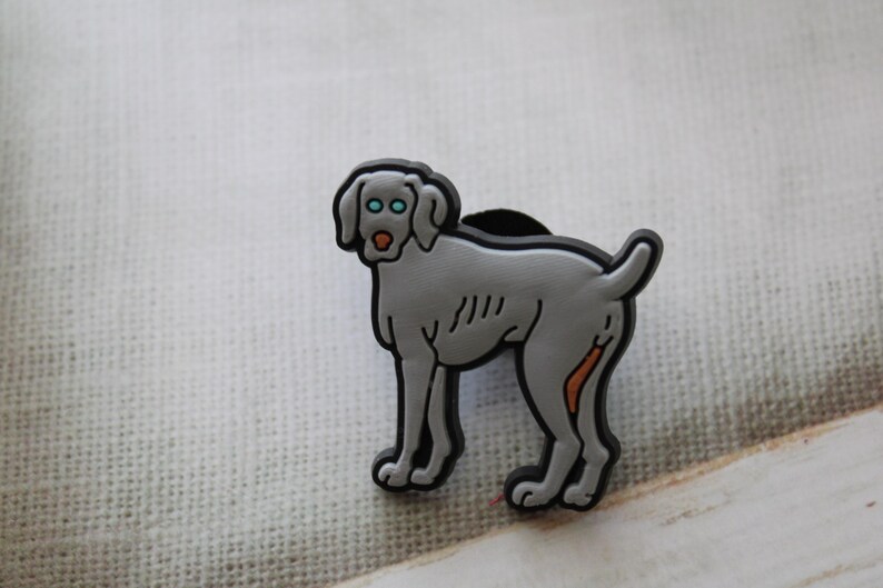 May include: A gray Weimaraner dog charm with blue eyes and an orange nose. The charm is made of rubber and has a black base.