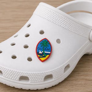 May include: A white foam clog shoe featuring a series of circular ventilation holes across the top and sides. Attached to the upper surface is a small, oval-shaped charm displaying the Guam seal, which includes a palm tree, a boat, and the text GUAM.