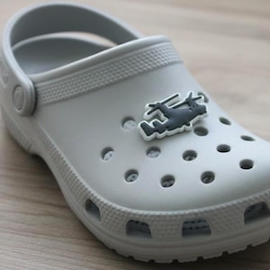 May include: A light gray clog shoe with a helicopter-shaped charm. The shoe has multiple ventilation holes and a pivoting heel strap. The charm is dark gray with a white outline. The shoe is made of a rubber-like material.