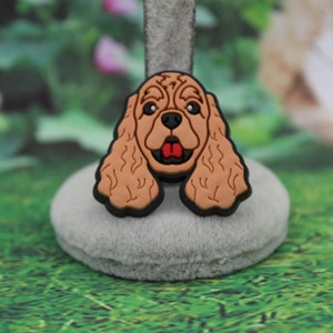 May include: A brown and black dog-shaped charm with a red tongue, set against a blurred green and white background. The charm is a detailed depiction of a Cocker Spaniel's face, with a black outline.