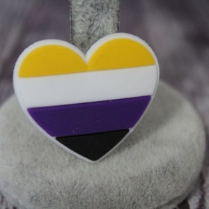 May include: A heart-shaped pin with the colors of the non-binary pride flag. The flag is yellow, white, purple, and black.