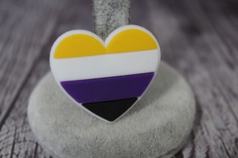 May include: A heart-shaped pin with the colors of the non-binary pride flag. The flag is yellow, white, purple, and black.