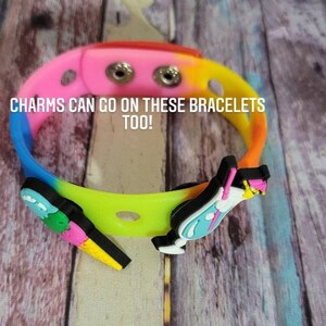 May include: A multi-colored silicone bracelet with two holes for charms. The bracelet has a rainbow gradient of pink, orange, yellow, green, blue, and purple. The text "CHARMS CAN GO ON THESE BRACELETS TOO!" is printed on the bracelet.