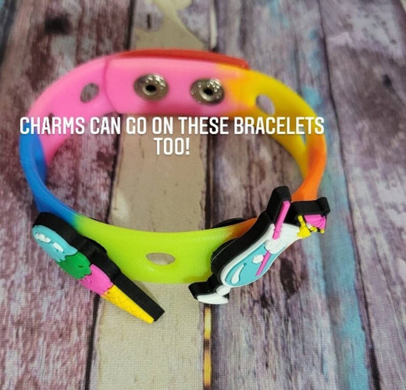 May include: A multi-colored silicone bracelet with two holes for charms. The bracelet has a rainbow gradient of pink, orange, yellow, green, blue, and purple. The text "CHARMS CAN GO ON THESE BRACELETS TOO!" is printed on the bracelet.