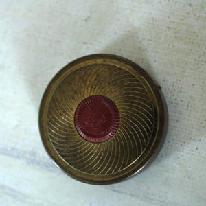 May include: A round, metallic gold-toned compact case featuring a textured, swirling radial pattern across its surface. The center of the lid is accented with a circular, deep red inlay that displays a small, embossed emblem or logo design.