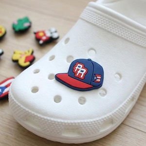 May include: A white foam clog shoe featuring a blue and red baseball cap charm with the letters PR and a small Puerto Rican flag detail. Several other colorful decorative charms are scattered nearby on the surface, including a flag design.