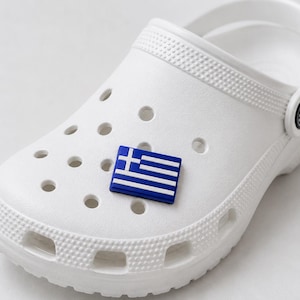 May include: A white foam clog shoe featuring a rectangular shoe charm shaped like the national flag of Greece. The flag charm is blue and white, displaying the characteristic cross and horizontal stripes, securely inserted into one of the ventilation holes.