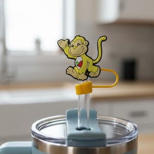 May include: A pale blue insulated travel mug with a stainless steel rim and handle. A yellow monkey straw topper with a red and white heart design sits on top of the clear straw. The mug is designed to keep drinks cold or hot.