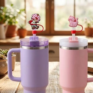 May include: Two insulated travel mugs, one lavender and one pink, with handles and lids. Each mug has a decorative straw topper, one with a Strawberry Shortcake character and the other with a pink cat. The mugs are on a wooden table.