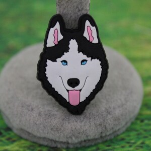 May include: A decorative shoe charm featuring a stylized husky dog head with black and white fur, bright blue eyes, pink inner ears, and a pink tongue hanging out. The design is rendered in a flat, graphic style with a black border outline.