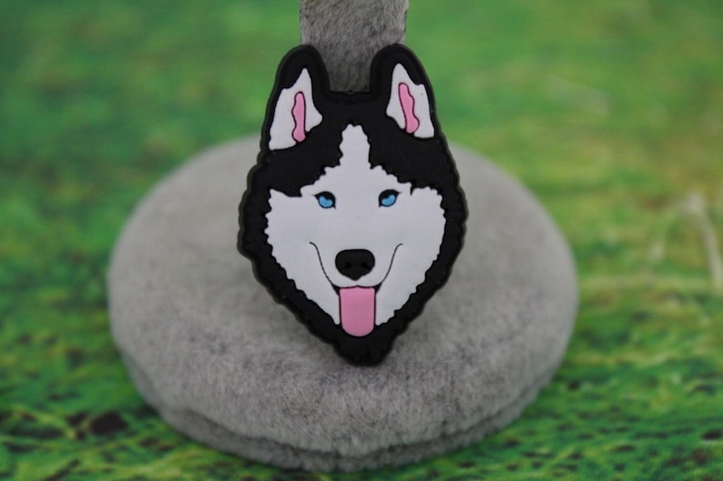 May include: A decorative shoe charm featuring a stylized husky dog head with black and white fur, bright blue eyes, pink inner ears, and a pink tongue hanging out. The design is rendered in a flat, graphic style with a black border outline.