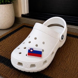 May include: A white foam clog shoe featuring a rectangular charm of the Slovenian flag attached to the top. The shoe has a rounded toe, multiple ventilation holes across the upper surface, and a pivoting heel strap with a circular black logo button.