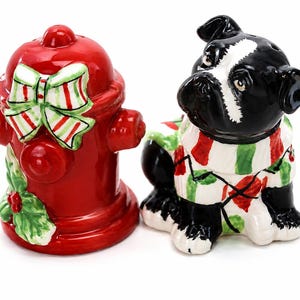 May include: A ceramic salt and pepper shaker set featuring a bright red fire hydrant decorated with a green and white striped bow and holly leaves, alongside a black and white dog wearing a green, red, and white patterned sweater.