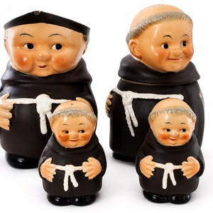 May include: A set of four ceramic monk figurines featuring two larger figures and two smaller figures. Each monk wears a dark brown habit with a white rope belt around the waist. The figures have painted facial features, rosy cheeks, and bald heads.