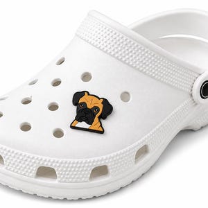 May include: A white foam clog shoe featuring a series of circular ventilation holes across the top and sides, with a decorative rubber charm shaped like a brown and black boxer dog head attached to one of the holes on the upper surface.