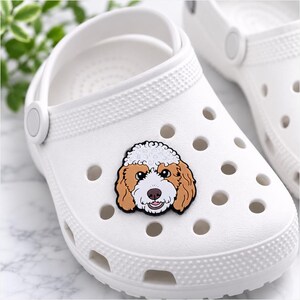 May include: A white foam clog shoe featuring a decorative charm shaped like a dog head with curly white and brown fur, dark eyes, and a pink tongue. The shoe has a rounded toe, a pivoting heel strap, and multiple circular ventilation holes.