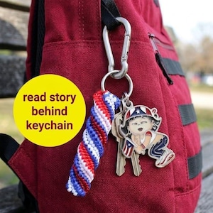 May include: A red backpack with a silver carabiner clip holding a keychain. The keychain has a red, white, and blue braided lanyard and a metal charm of a child in patriotic attire. The text on the yellow circle says "read story behind keychain".
