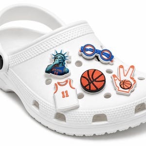 May include: A white clog shoe decorated with five rubber charms featuring a Statue of Liberty figure, blue glasses, a basketball, a white jersey with the number 11, and a hand gesture sign with a basketball, all attached to the shoe's ventilation holes.