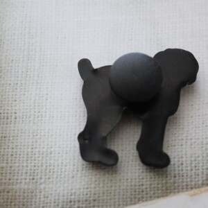 May include: Black metal silhouette of a dog with a magnetic back. The dog is standing on a white fabric surface.