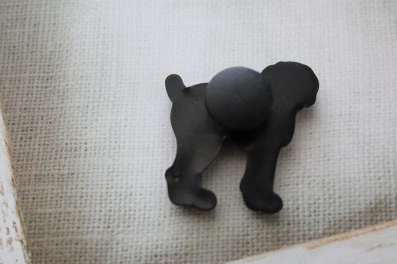 May include: Black metal silhouette of a dog with a magnetic back. The dog is standing on a white fabric surface.